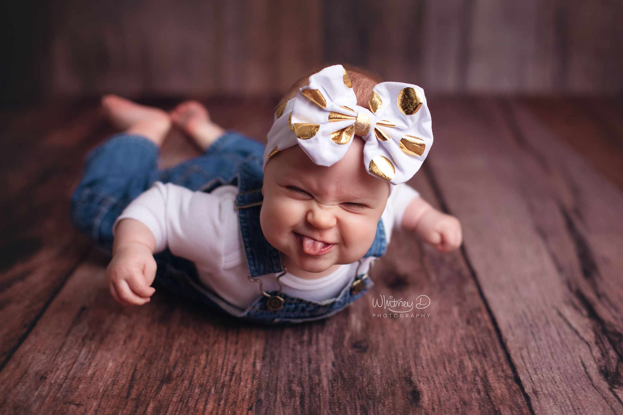 Six month old baby girl in overalls and big bow sticking her tongue out at Whitney D. Photography in Conway, Arkansas